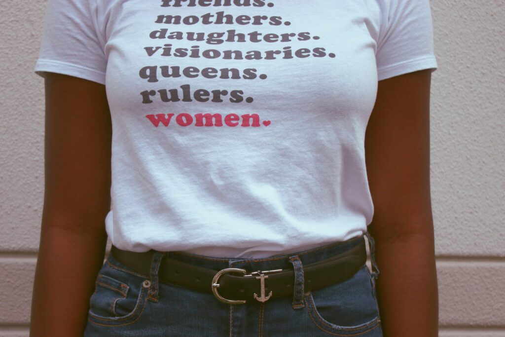 Schwarze Frau von Schultern bis zur Hüfte. Auf ihrem weißen T-Shirt steht: friends. mothers. daughters. visionaries. queens. rulers. (braun) women. (rot)